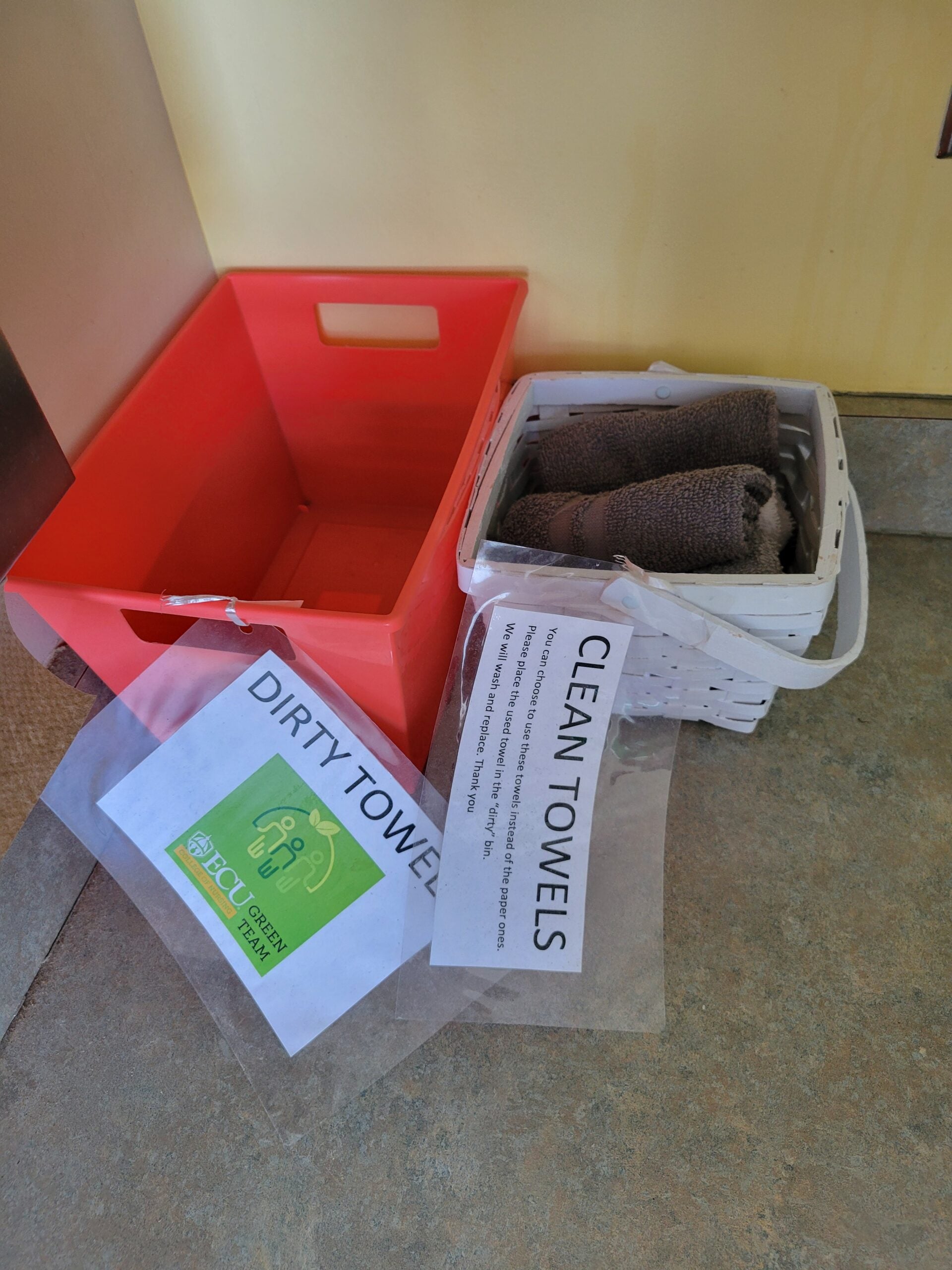 A basket filled with clean hand towels labeled "Clean Towels" next to an empty bucket labeled "Dirty Towels"