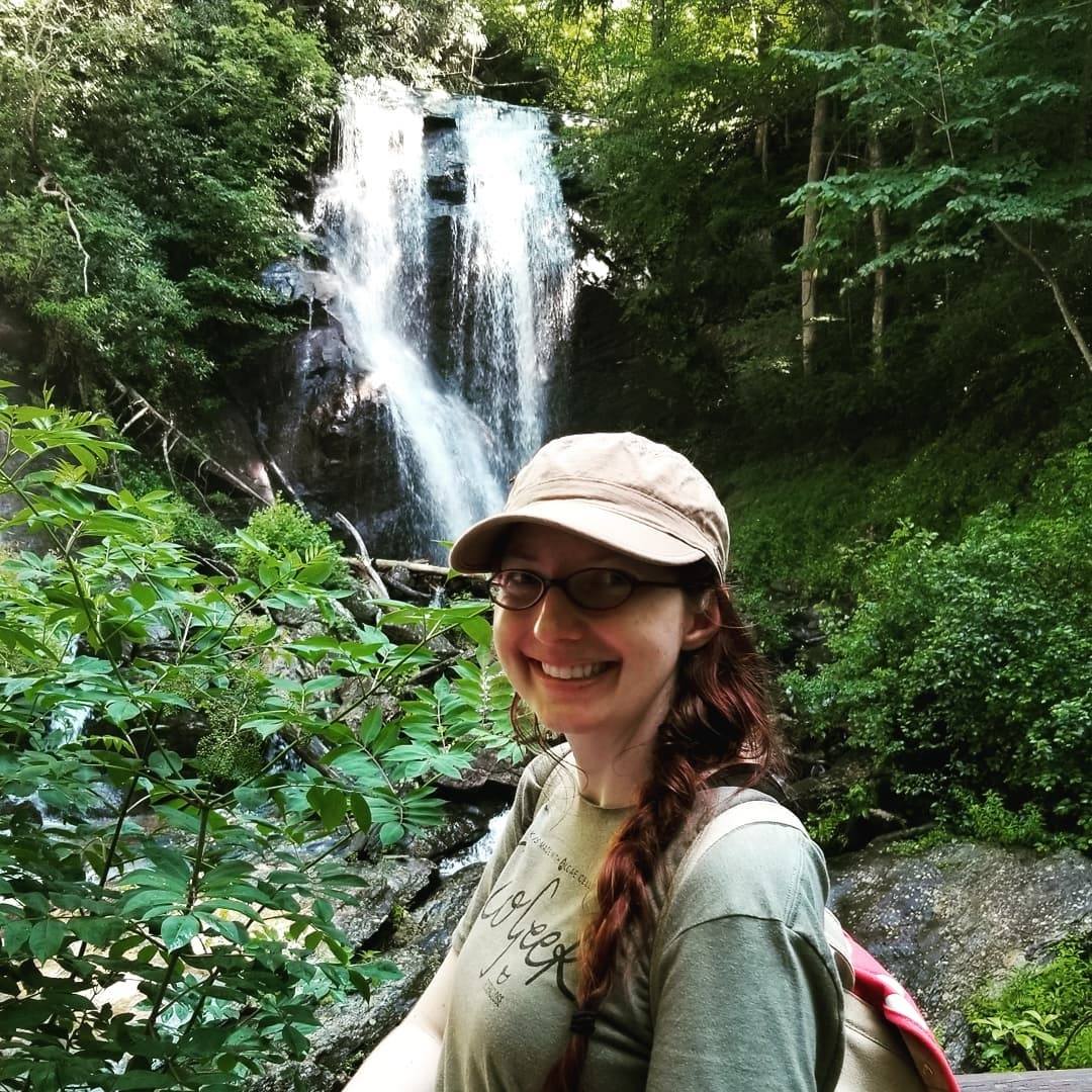 Kim Fox, a woman with long braided hair in a hat, stand in front of a waterfall