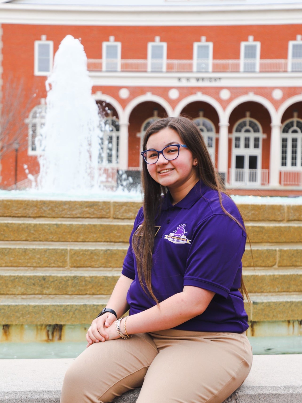 Tori Bain, a young woman with long hair and glasses, sits in front of ECU's fountain