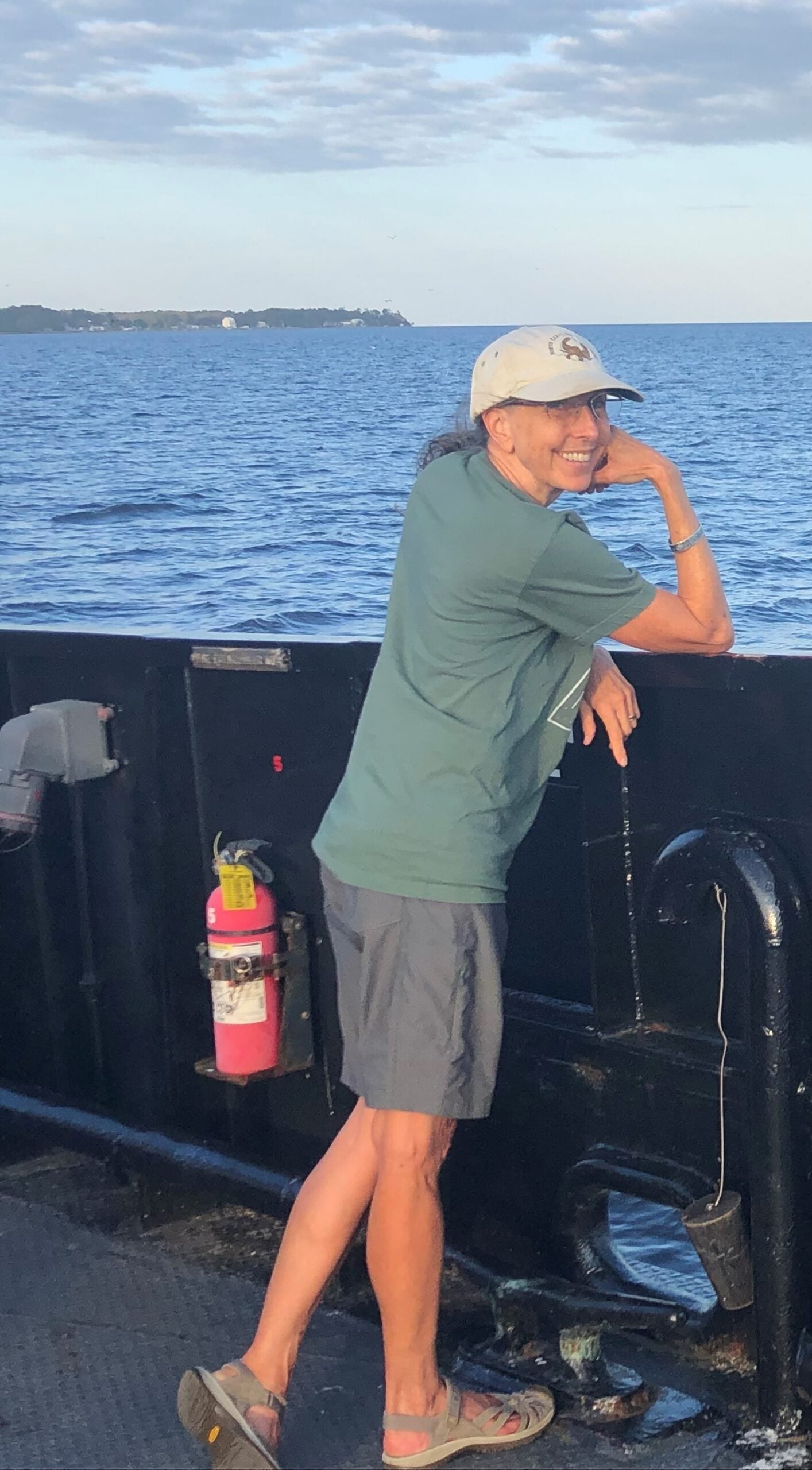 L.J. Palmer-Maloney, a woman with gray hair in a ponytail and a baseball cap, looking back over her shoulder with a smile while standing by the ocean