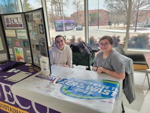 Two young women sit smiling at a table with information about sustainability and a banner for Campus Race to Zero Waste, collecting signatures