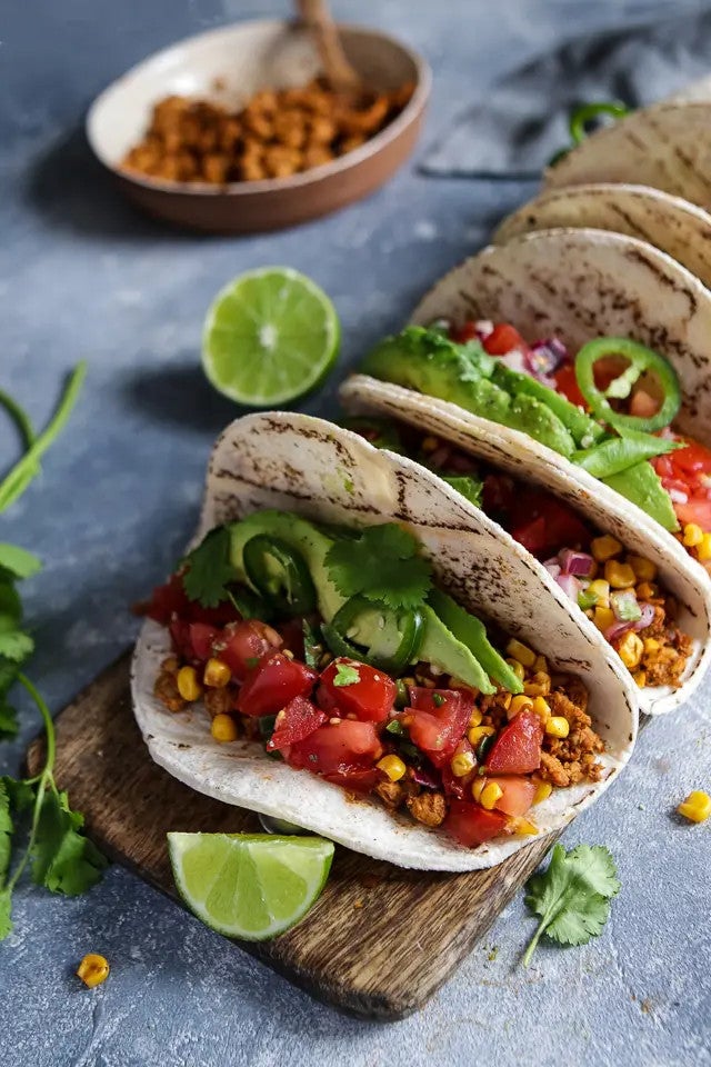 Three vegan tacos are lined up on a plate with tempeh, corn, salsa, and lettuce