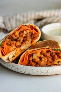 A buffalo chickpea wrap cut in half and sitting on a plate next to a white cup of white sauce.