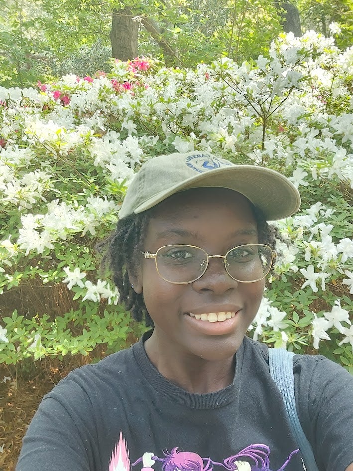 A young woman in a baseball cap smiles at the camera with a white flower bush in the background