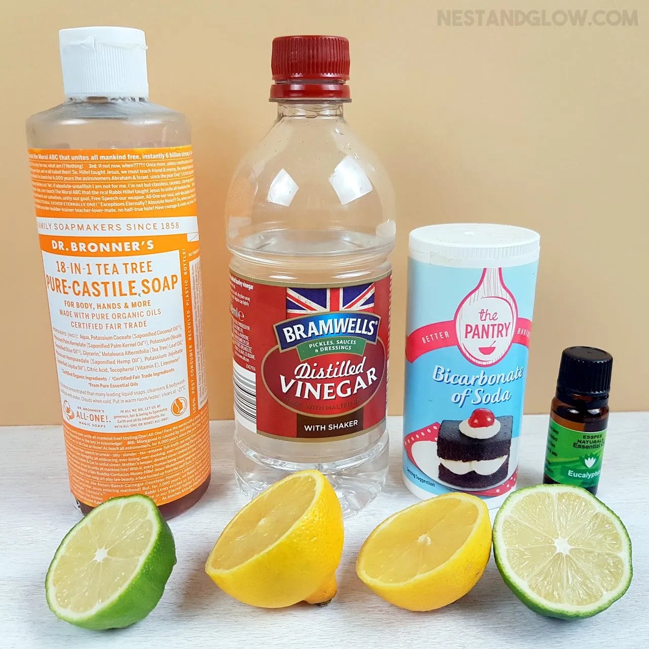 4 bottles lined up in a row: castile soap, distilled vinegar, bicarbonate soda (baking soda), and essential oil, all behind 2 halves of a lime and 2 halves of a lemon, also in a row